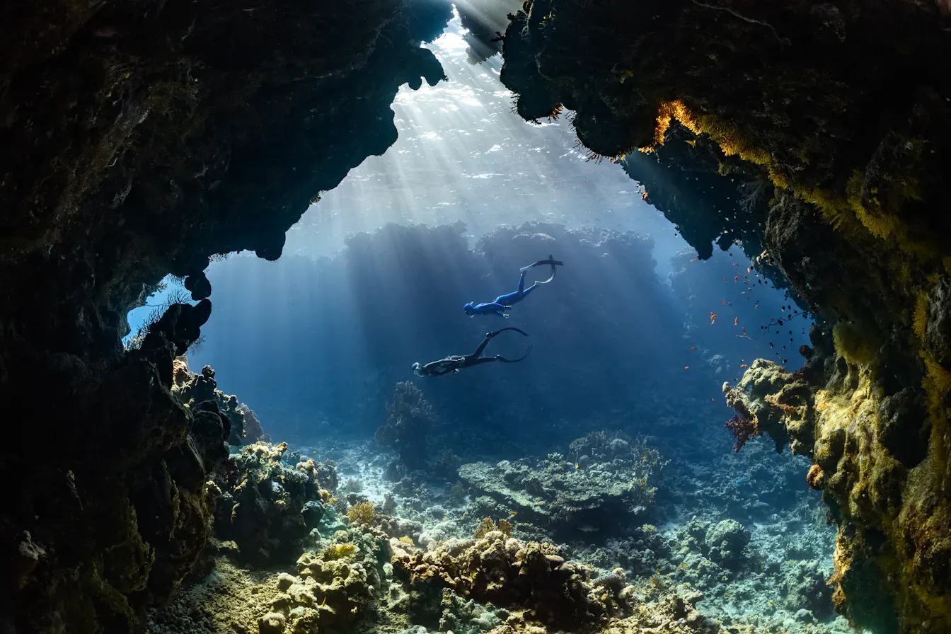Two freedivers descending together through a sunlit underwater cavern